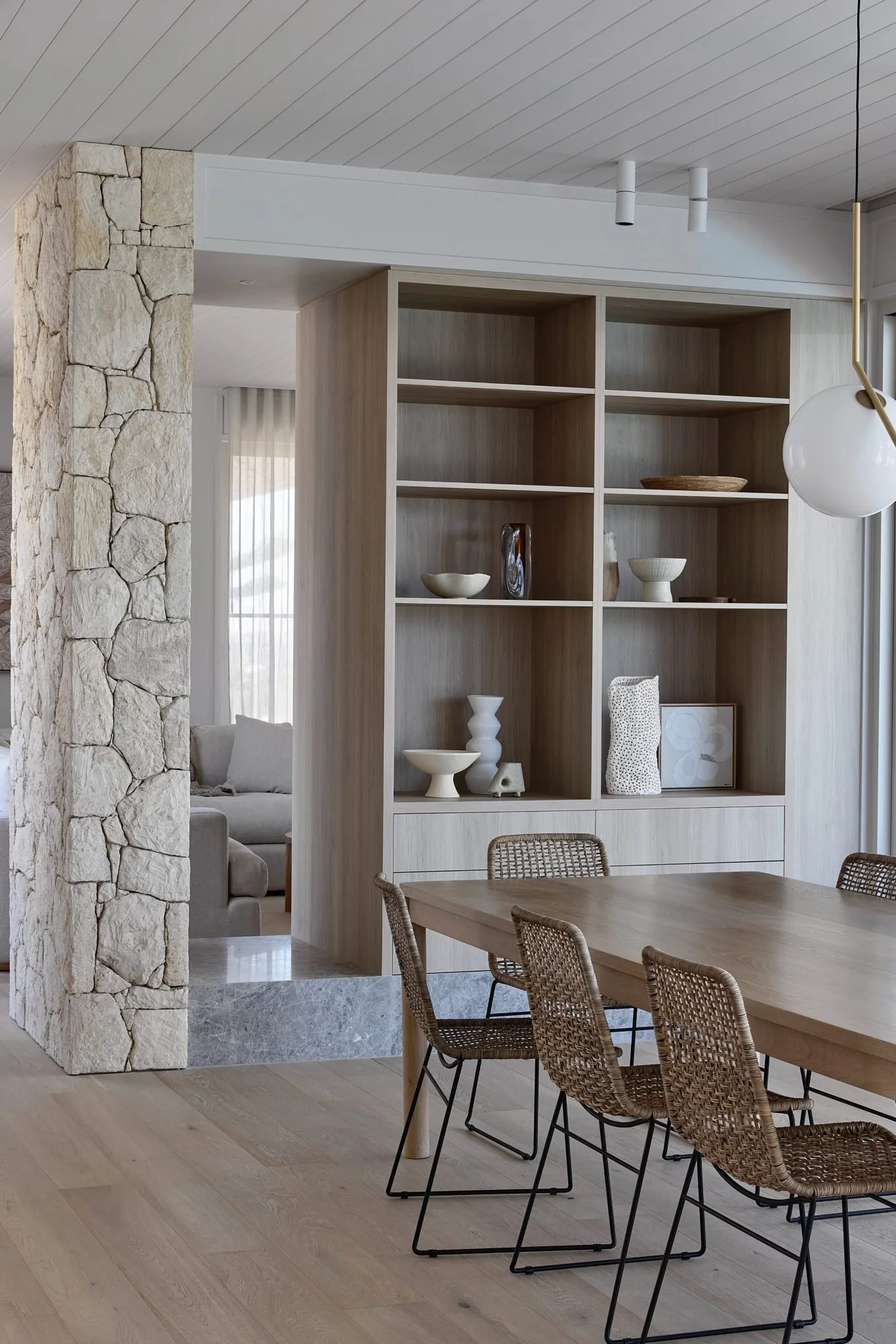 A minimalist dining room with a wooden dining table, six woven rattan chairs, and a built-in wooden shelf with decorative items. There is a stone column and a partial view of a living area with a Couch and curtains in the background.