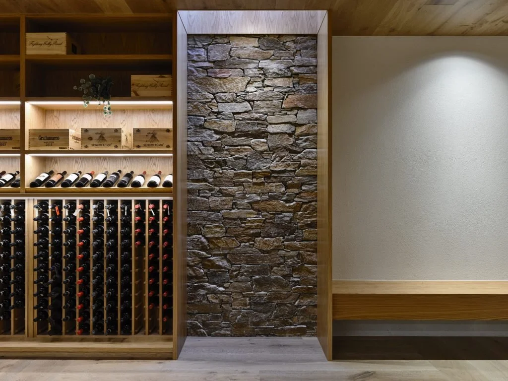 Wine storage area with wine bottles on wooden racks, a stone wall in the center, and a bench to the right.