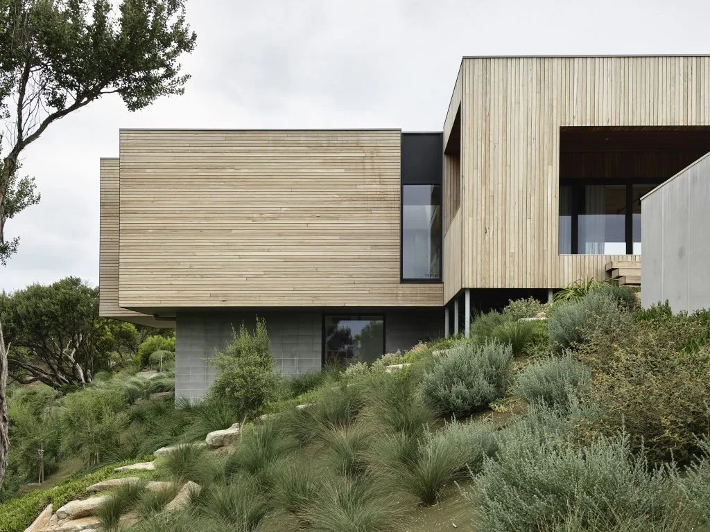 Modern house with wooden exterior, large windows, and landscaped hillside garden in daytime.