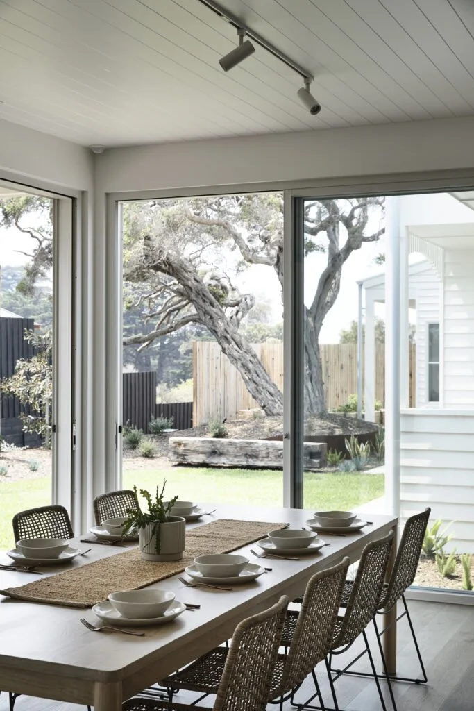 Dining room with a large glass sliding door, view of a backyard with trees and green grass, six set plates and silverware on the table, potted plant in the center.