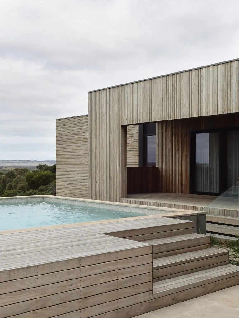 Modern wooden house with a swimming pool on a deck, overlooking a green landscape under a cloudy sky.