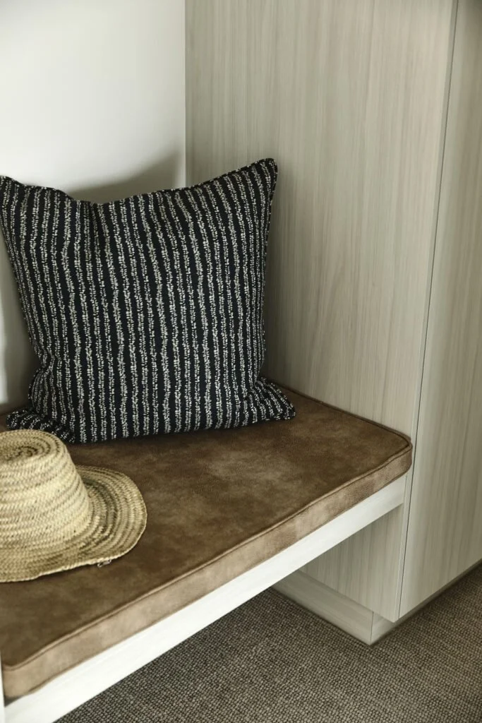 A brown cushion on a built-in bench with a black and white striped pillow and a straw hat placed on the bench.
