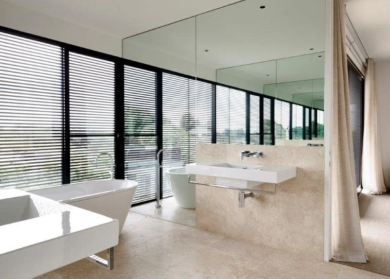 Modern bathroom with large windows, black blinds, white bathtub, and a rectangular sink with a mirror above.