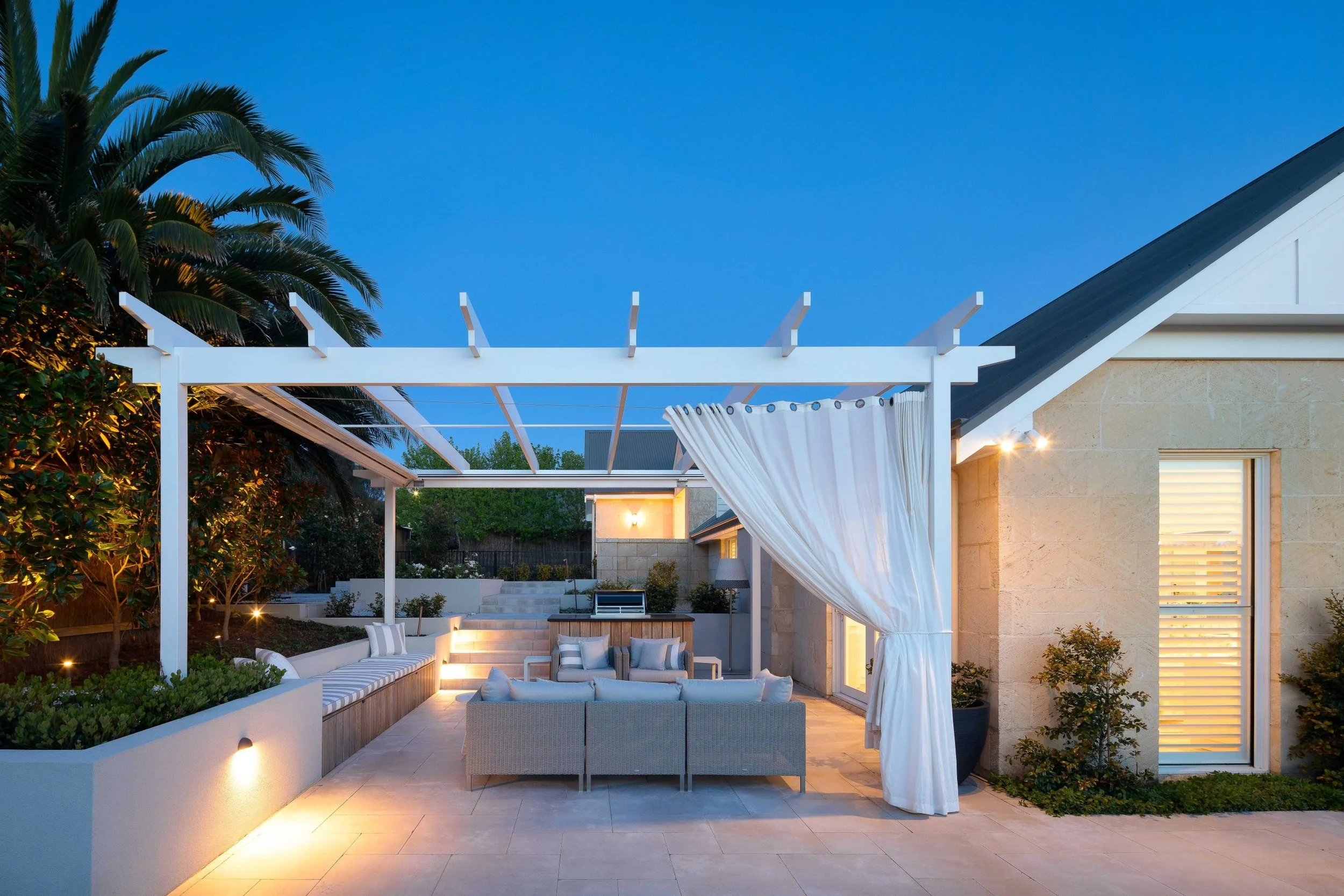 Elegant outdoor patio with comfortable seating, white curtains, and ambient lighting, surrounded by greenery and a modern house with a sloped roof. Palm trees are visible on the left side during dusk.