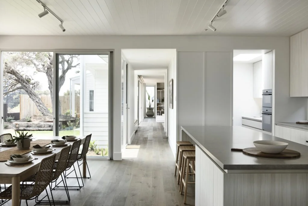 Bright, modern kitchen and dining area with large window overlooking a garden, white cabinetry, and a spacious island with wooden stools.