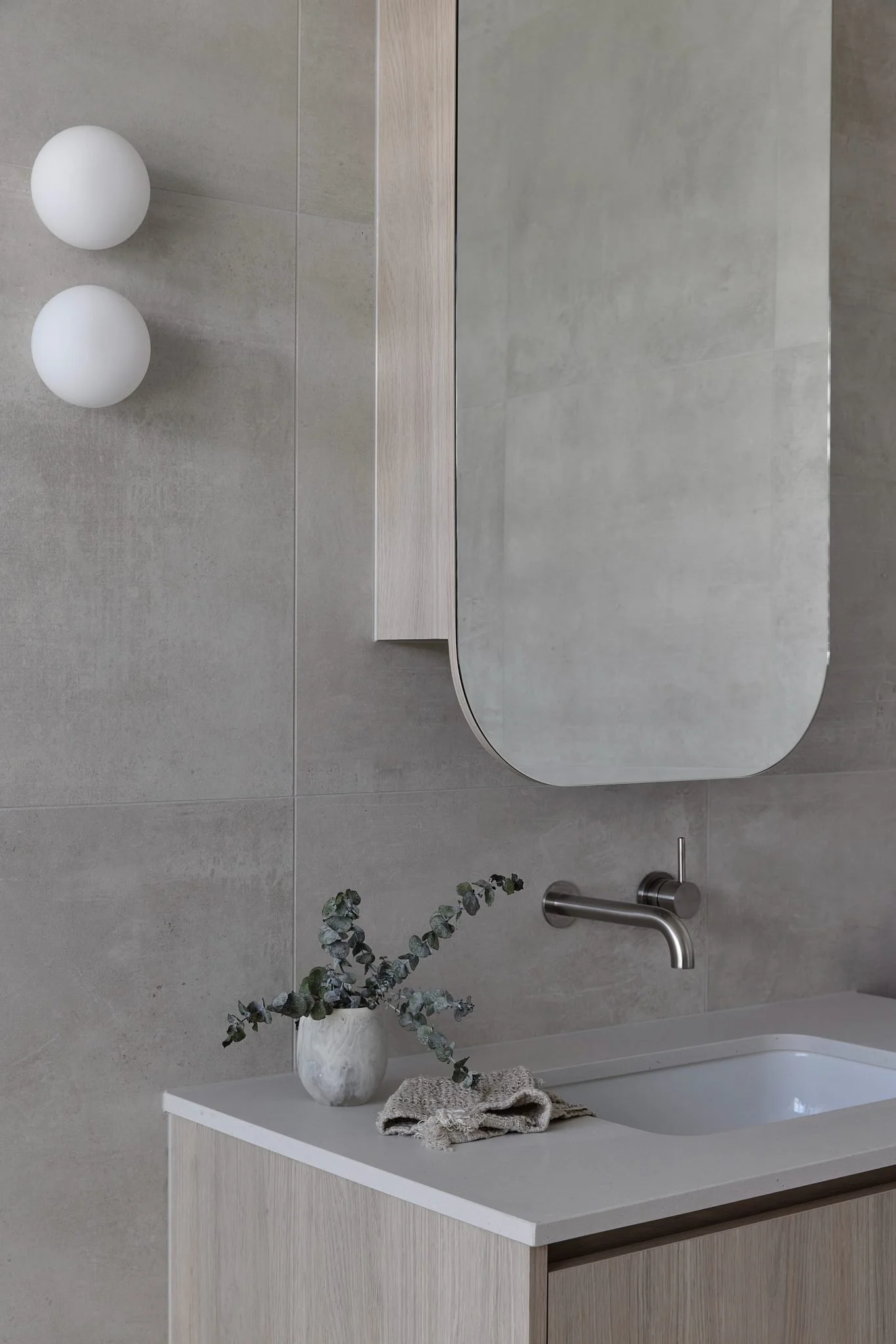 A minimalist bathroom with a wall-mounted mirror, a modern faucet, a small plant in a white pot, a folded towel, and two round white wall sconces.