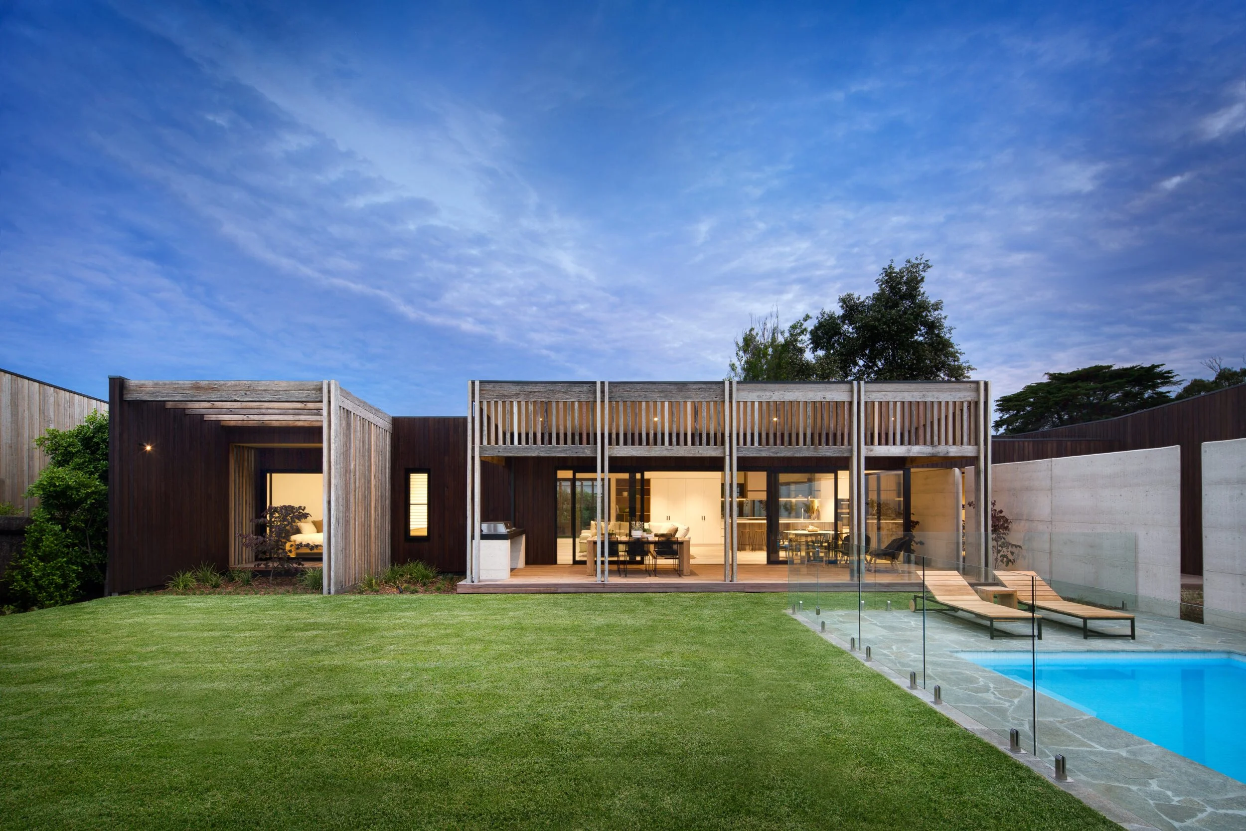 Modern house with large glass sliding doors, a swimming pool with two lounge chairs, a well-maintained green lawn, and a blue sky with clouds.