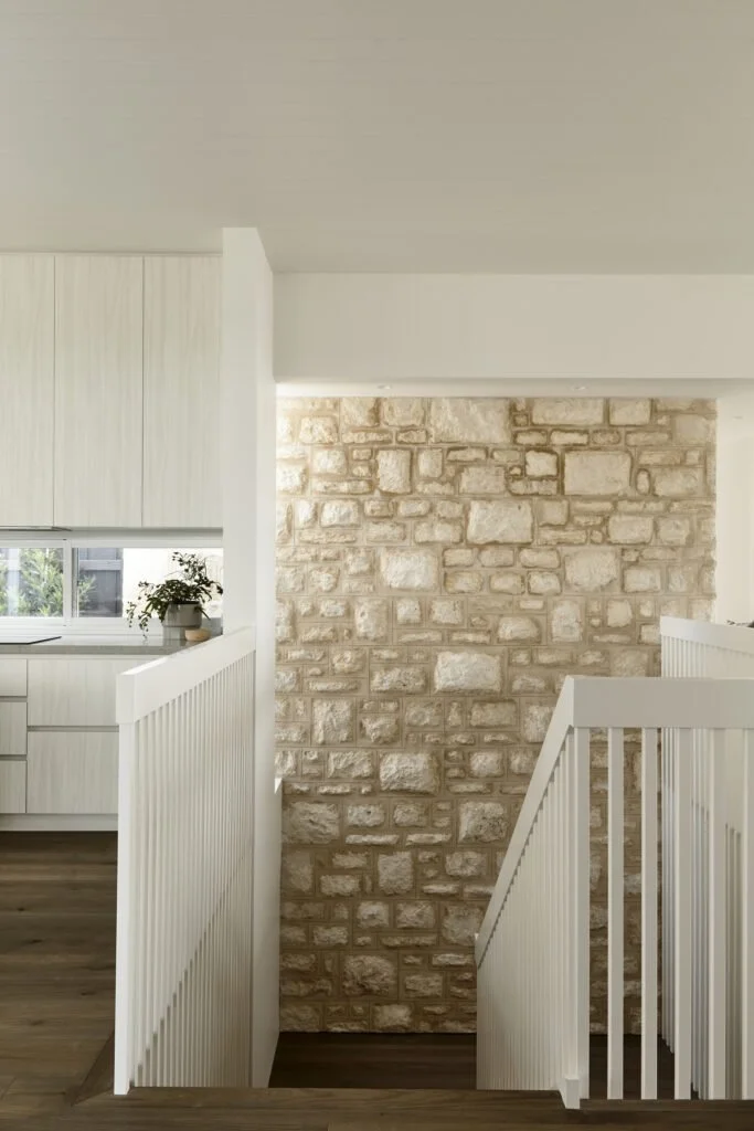 Interior view of a staircase with white railing leading down to a lower level, featuring a stone accent wall, part of a kitchen with white cabinets and a window with a plant.