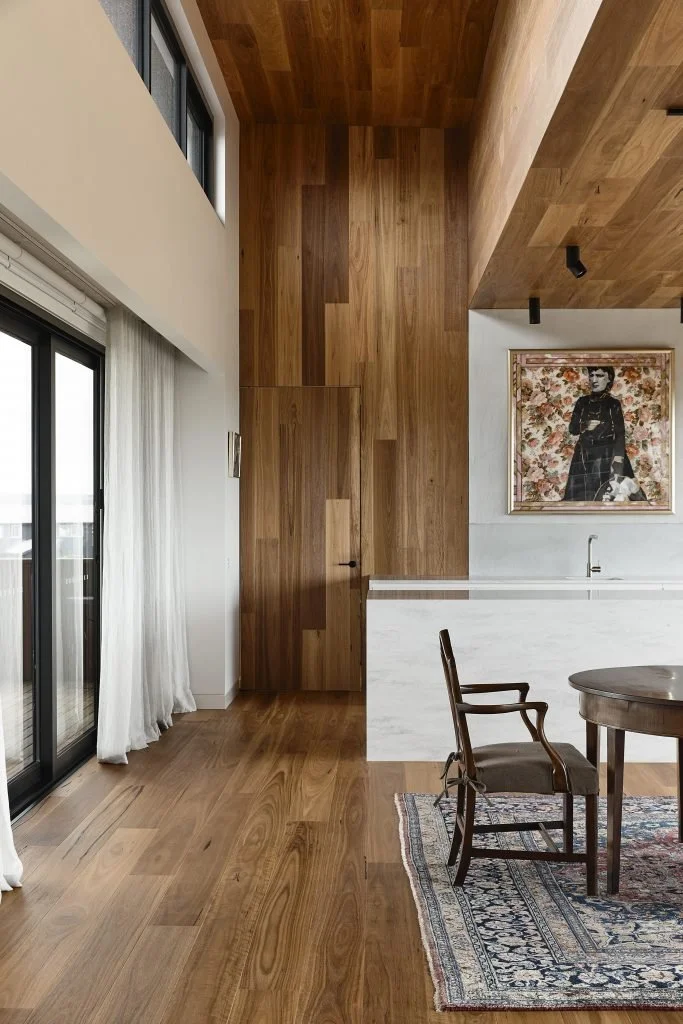 Interior view of a modern apartment with wooden flooring and walls, a sliding glass door with white curtains, a wooden chair and round table on a patterned rug, a white kitchen countertop, and a framed artwork of a woman in black on the wall.