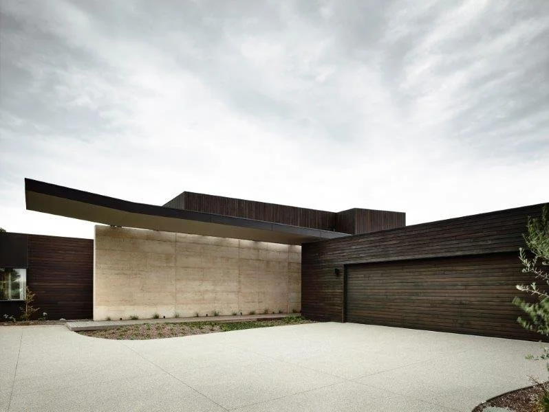 Modern house with a flat roof, accented by a large overhang, featuring dark wood and concrete exterior walls. The driveway in front is paved, and there's a small garden area with minimal landscaping.
