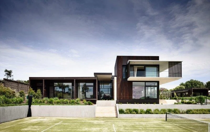 Modern multi-story house with large windows and balconies, situated on a grassy yard with a tennis court in the foreground, under a partly cloudy sky.