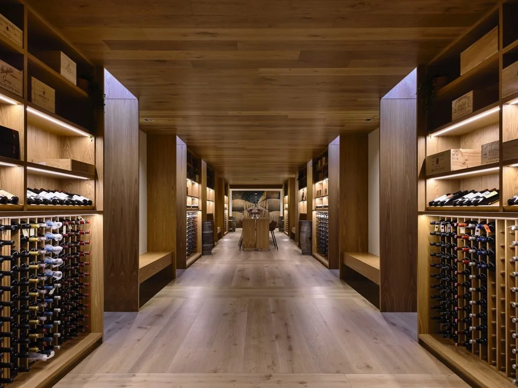 A wine cellar with wooden shelves stocked with bottles, illuminated with warm lighting, and a central table with chairs.