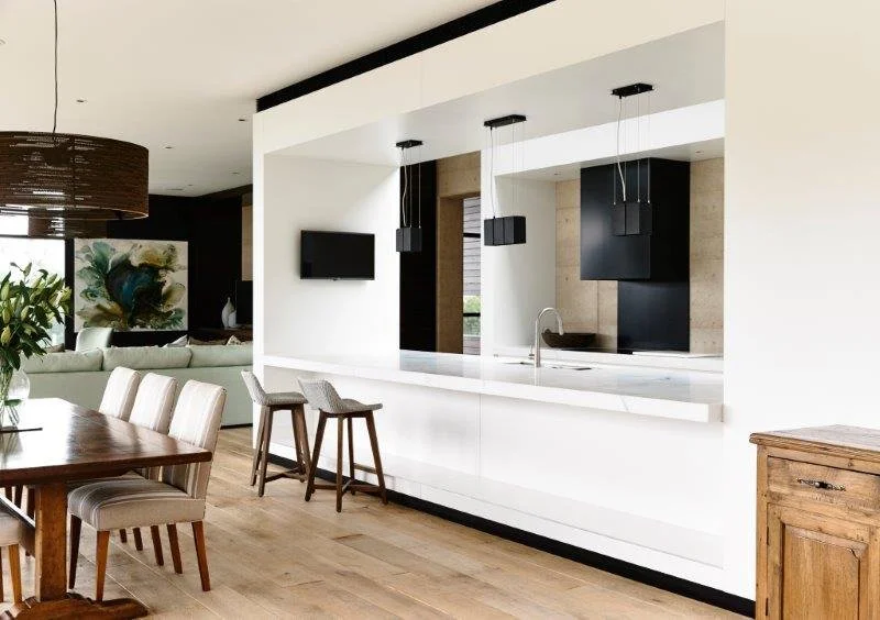 Open-concept living space with a kitchen and dining area, modern white kitchen with black accents, bar stools, wooden dining table with chairs, and a living room with a large abstract painting and greenery.