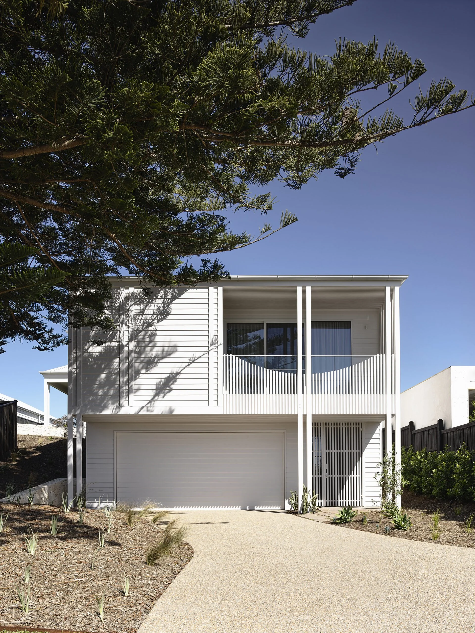 Modern white two-story house with a garage and a balcony, surrounded by plants and trees, under a clear blue sky.