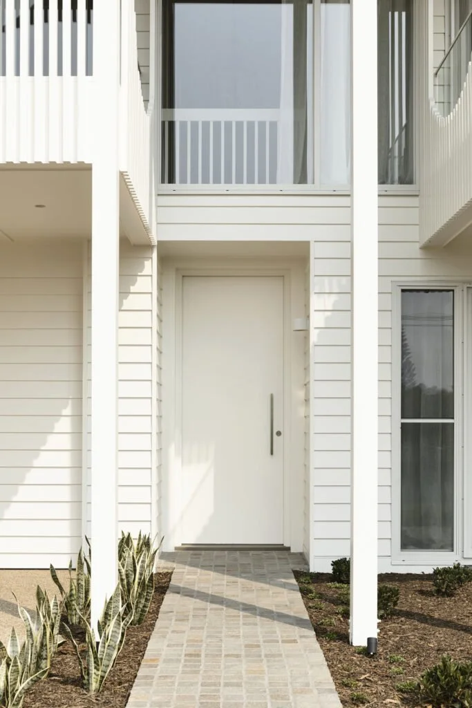 Modern white apartment building with a central entrance door, a brick pathway leading to it, and small plants on either side.