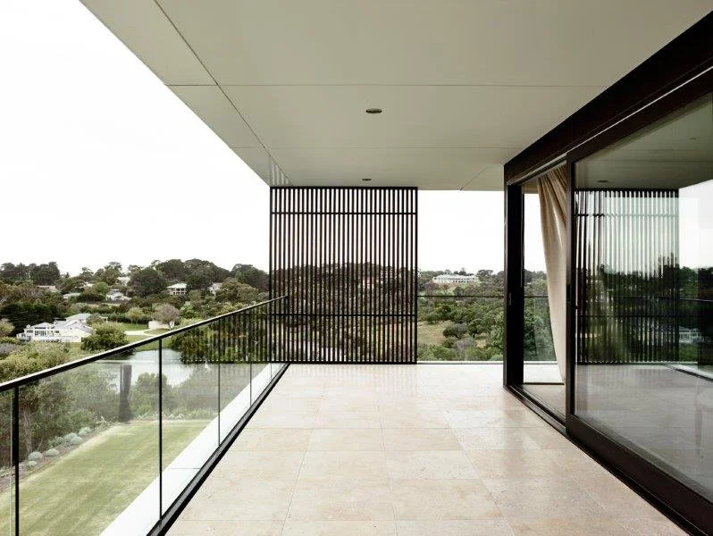 Modern balcony with glass railing, sliding glass door, and wooden privacy screen overlooking trees and suburban houses.
