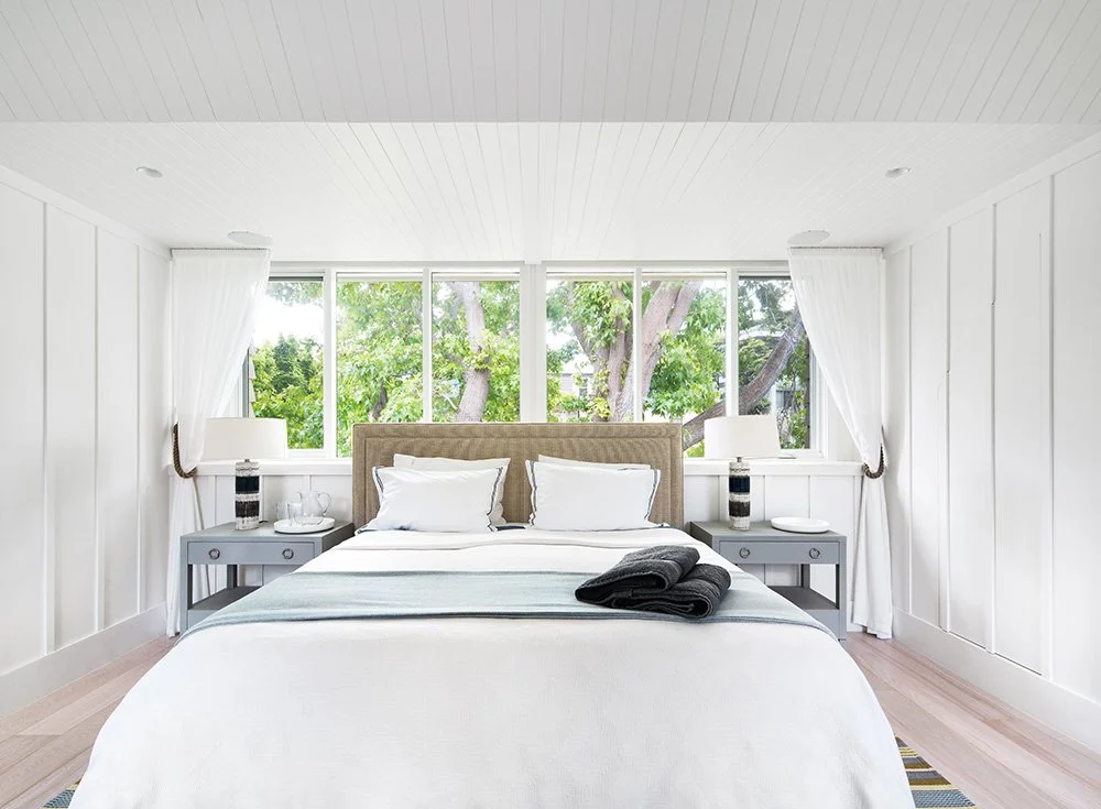 Bedroom with white walls, large window showing trees outside, bed with beige headboard, white bedding, black towels, nightstands with lamps, and curtains on both sides.