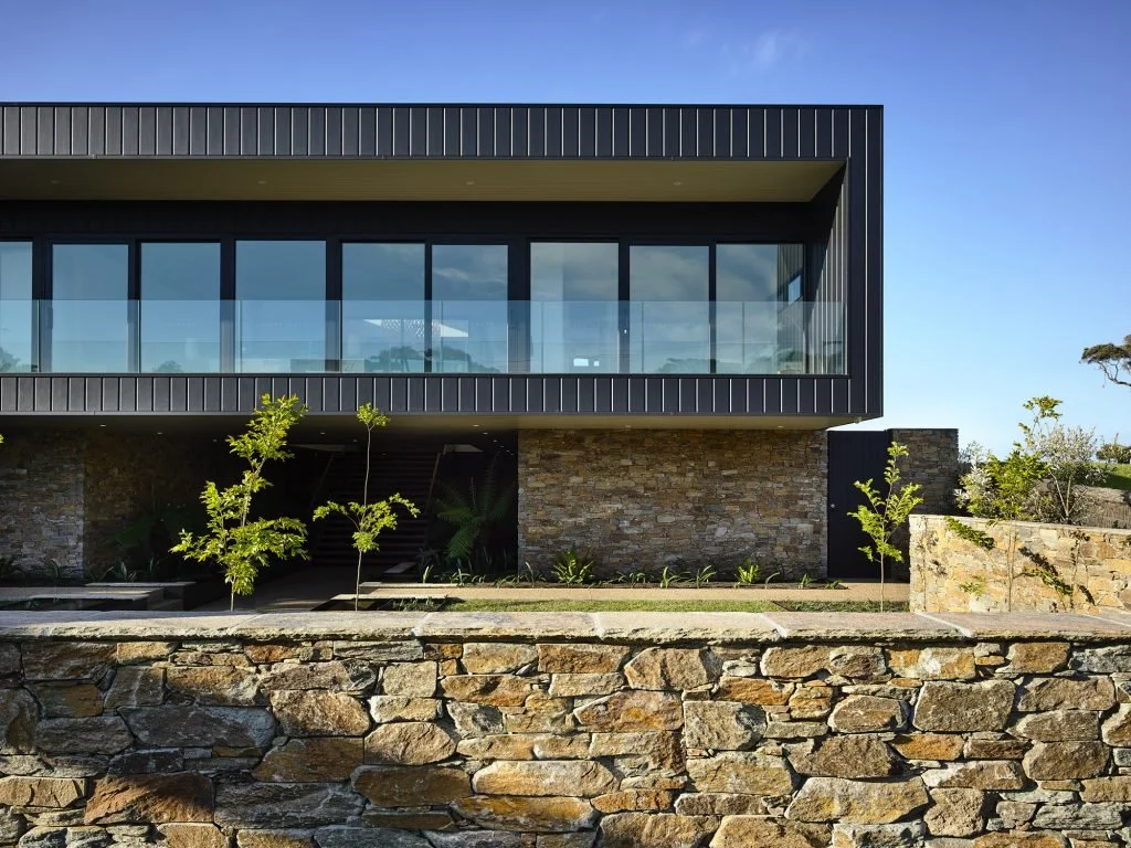 Modern house with large glass windows, stone walls, and a landscaped yard with small trees and a stone retaining wall under a blue sky.
