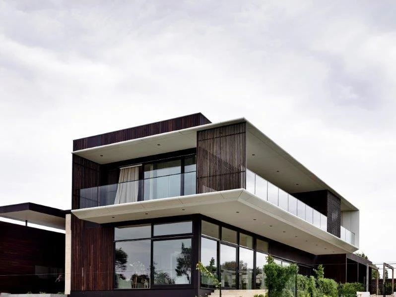 Modern multi-story house with large glass windows, dark wood paneling, and a balcony with glass railing under a cloudy sky.