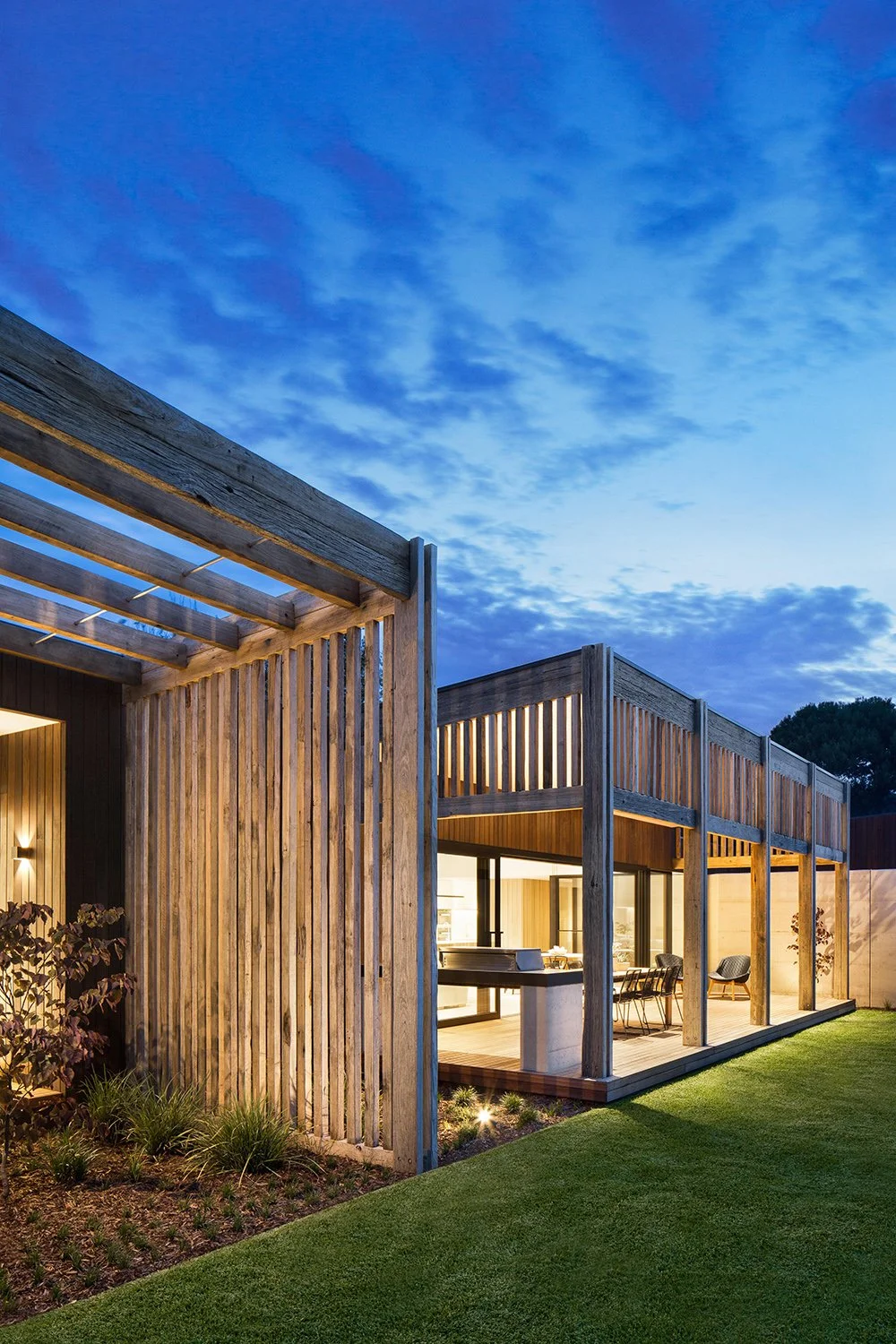 Modern house with wooden exterior and large windows, illuminated from inside, with a green lawn in the foreground and a blue sky at dusk.