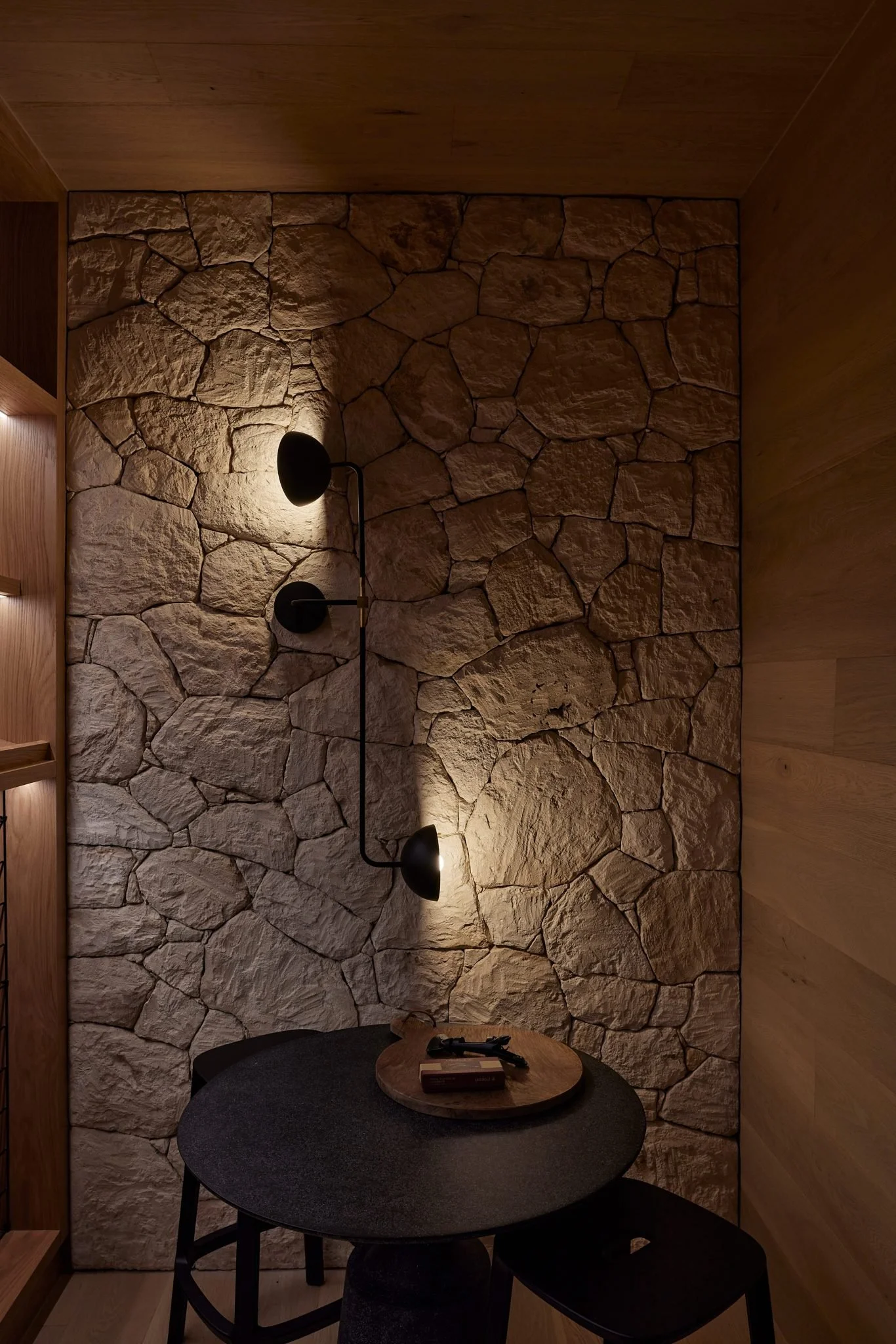 A cozy corner with a stone accent wall illuminated by a modern black wall-mounted light fixture, a round black table, and a wooden tray with a few small items.