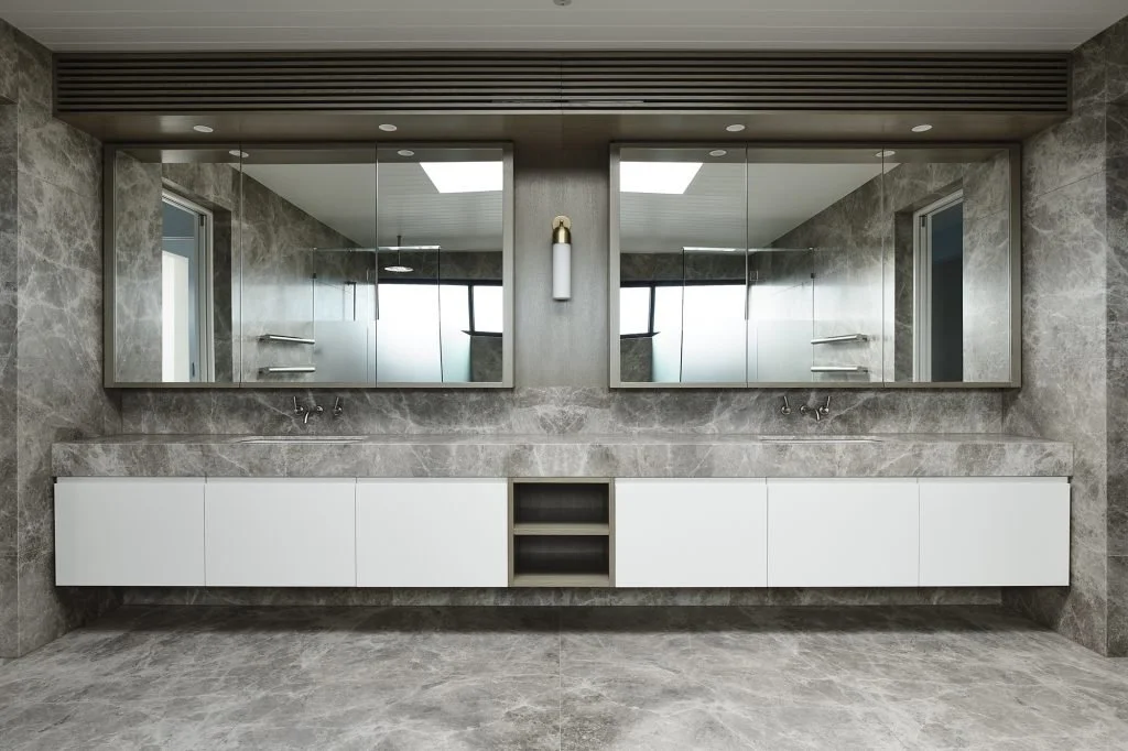 Modern bathroom with large mirrors, double sinks, and white storage cabinets against a marble wall and floor.