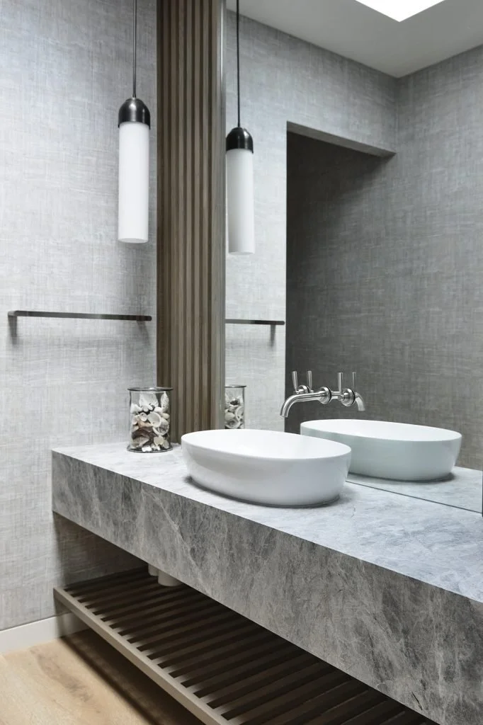 Modern bathroom with a gray marble countertop, two white vessel sinks, and a large mirror. Two pendant lights hang above the sinks, and decorative jars with shells are placed on the counter.