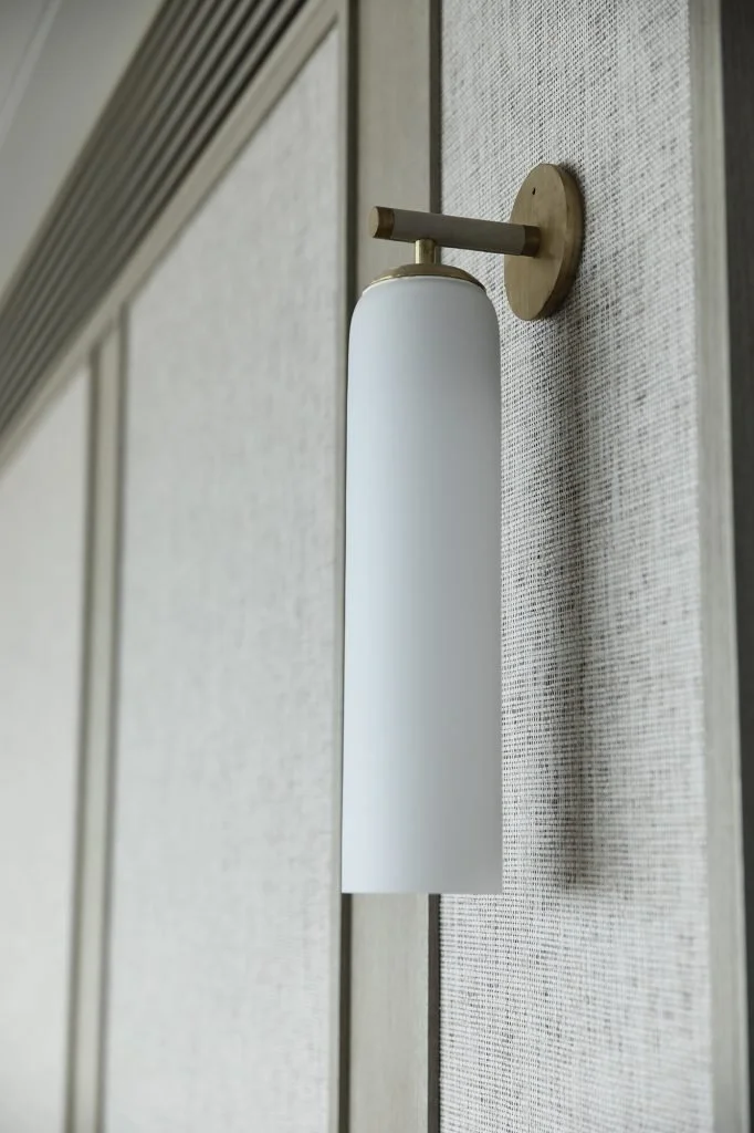 Close-up of a modern wall-mounted light fixture with a white cylindrical shade and a brass arm attached to a textured wall.