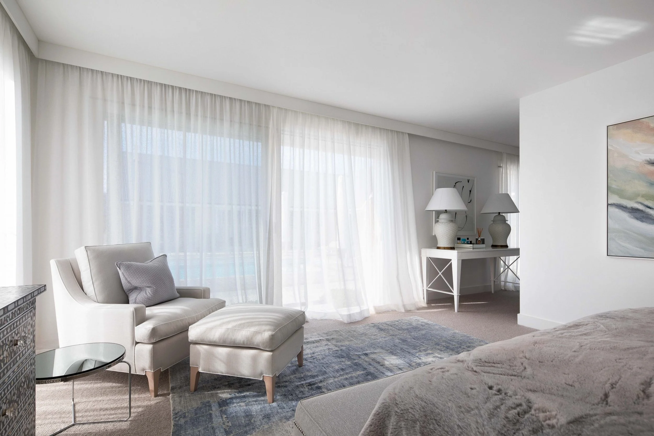 Bright hotel suite with white sofa, gray pillow, large window with sheer curtains, white table with lamps, and a bed with beige bedding.