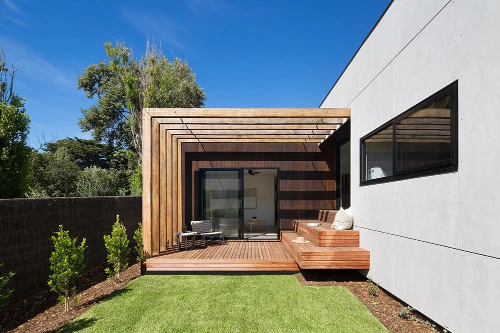 Modern backyard with concrete house wall, wooden deck, outdoor seating, and lush green lawn under a blue sky.