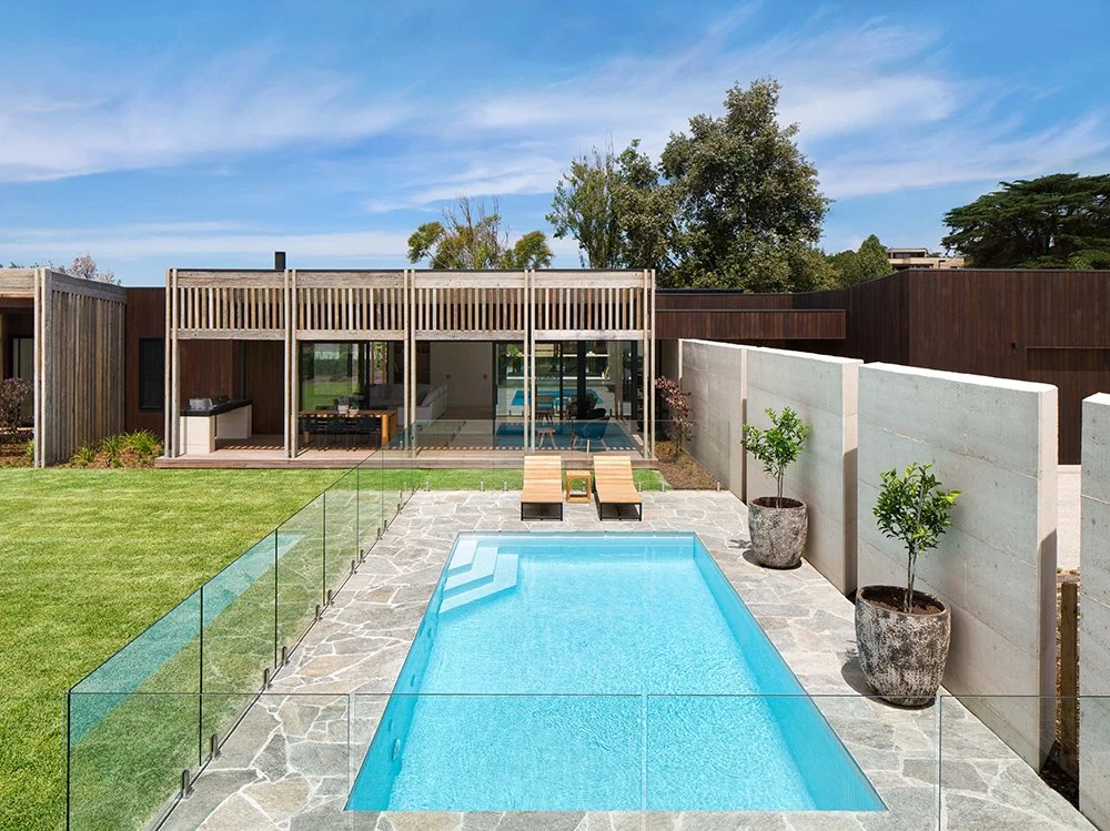 Modern backyard with rectangular swimming pool, glass safety fencing, two lounge chairs, potted plants, and a contemporary house with large glass doors and wooden exterior.