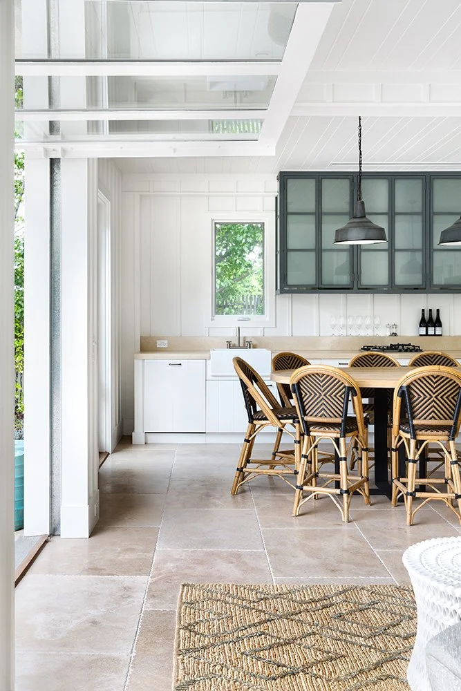 Bright modern kitchen with white walls and ceiling, beige tile flooring, a window, green cabinetry above the counter, a white farmhouse sink, and a wooden dining table with six wicker chairs. A patterned rug is partially visible in the foreground.