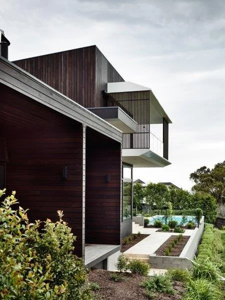 Modern residential building with dark wood exterior, large windows, and a balcony, surrounded by lush greenery and a landscaped yard with a pathway and swimming pool in the background.
