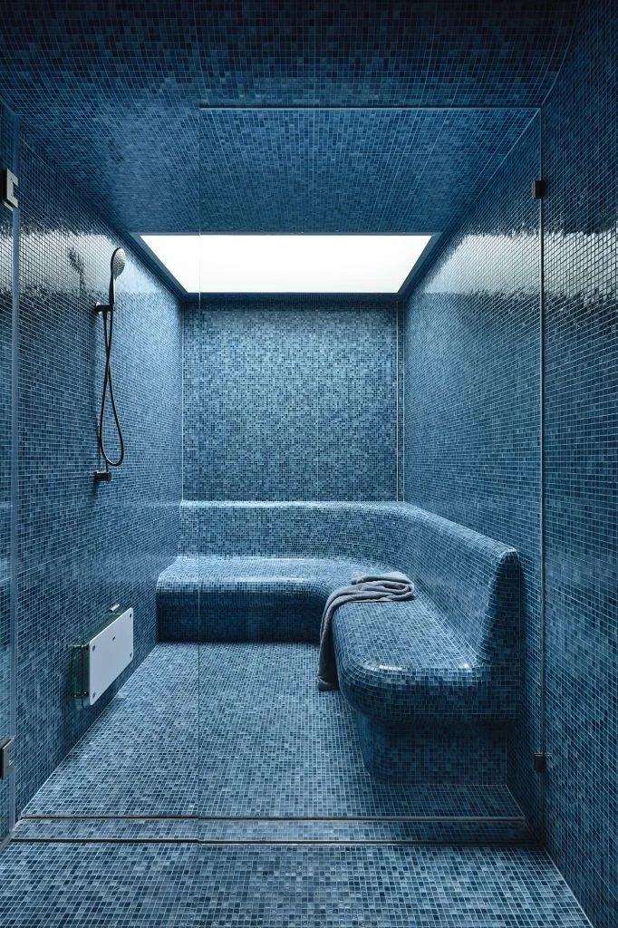 Blue mosaic-tiled steam room with a curved bench, handheld shower, and overhead skylight.