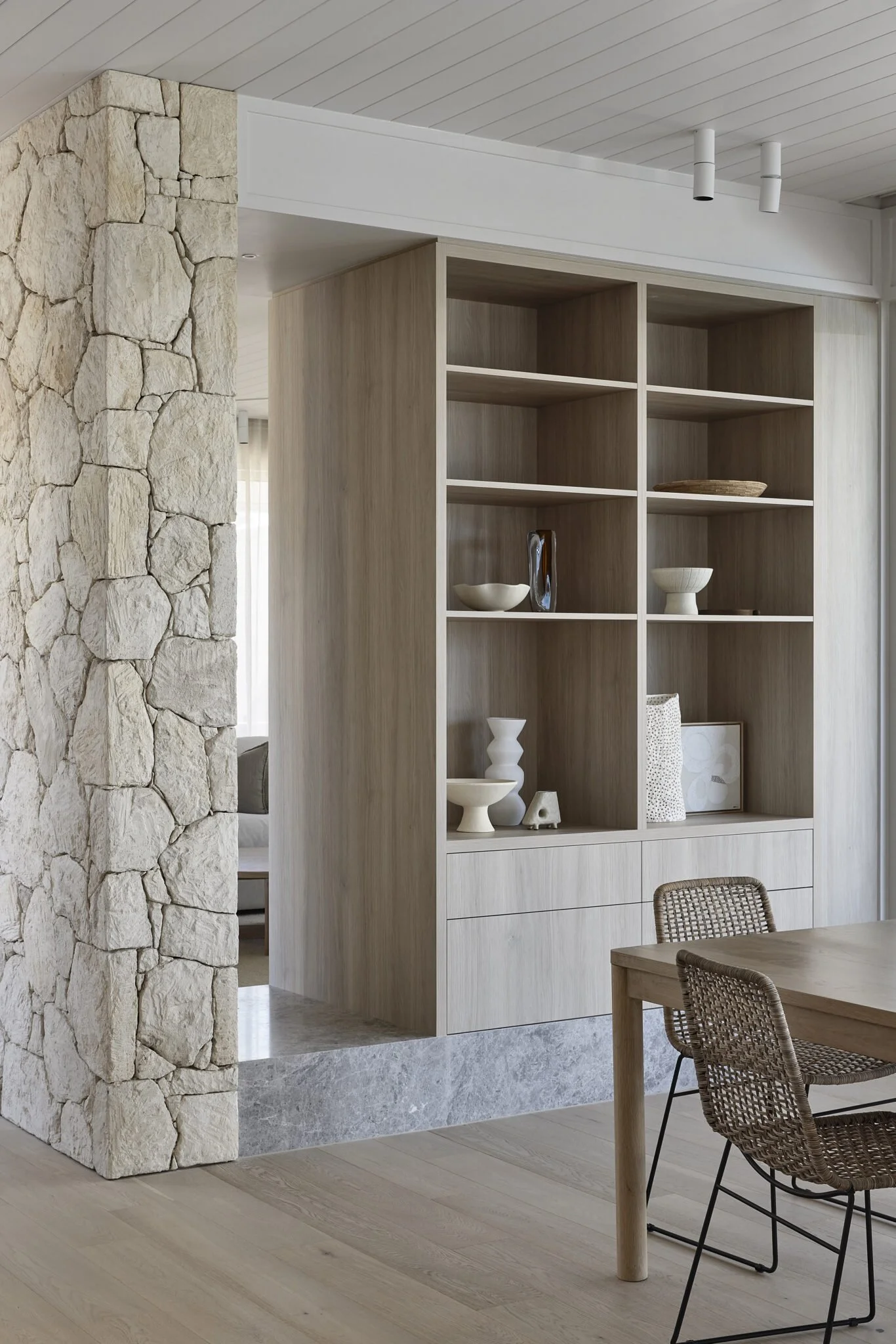 Corner of a dining area with a light wood built-in shelving unit displaying decorative objects, next to a stone wall and a wooden table with wicker chairs.