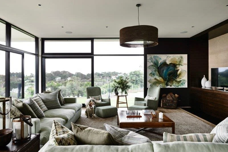 Modern living room with large windows, beige sofas, a green armchair, a dog, a painting, and a wooden coffee table.
