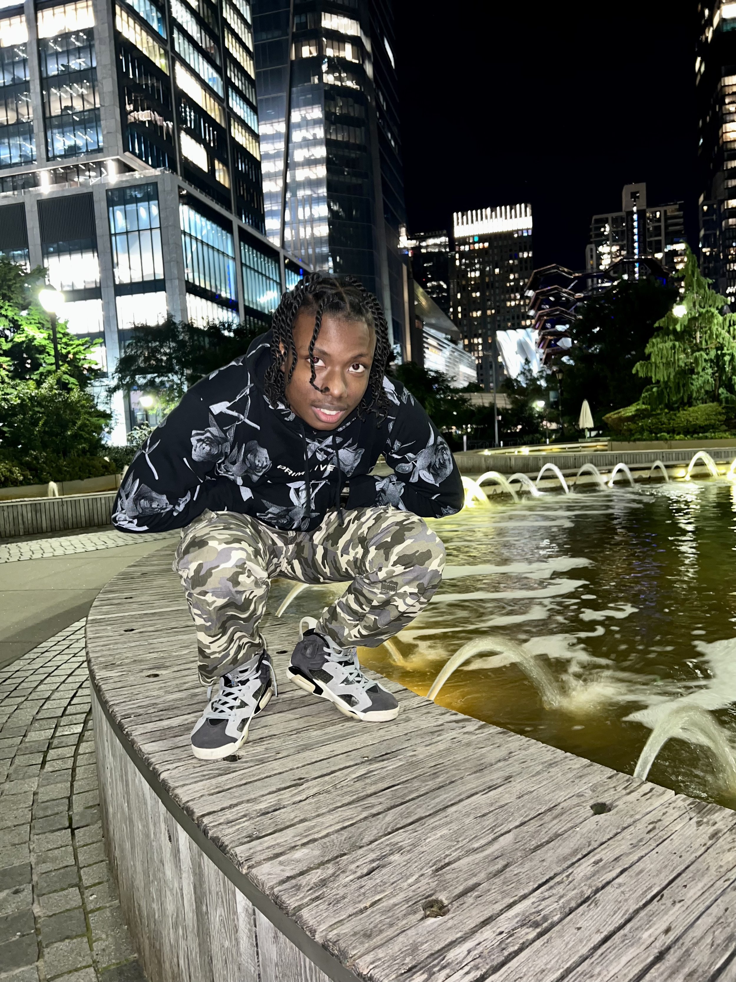 A person squatting by a fountain at night in an urban setting, with tall illuminated buildings in the background.