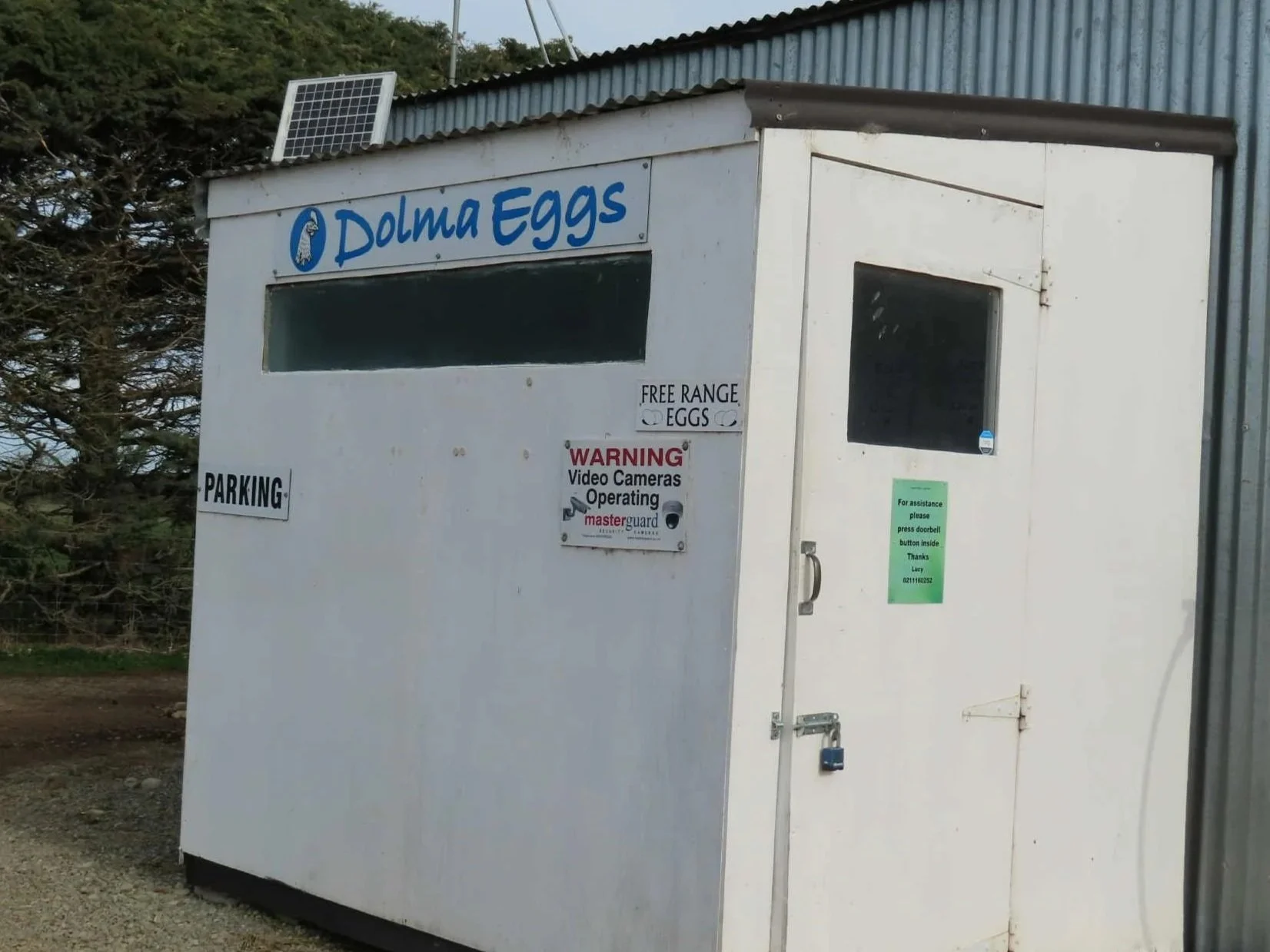 A small white structure with a sign reading "Dolma Eggs" on top, a solar panel on the roof, and windows. There are additional signs indicating free-range eggs, parking, and a warning about video cameras operating.