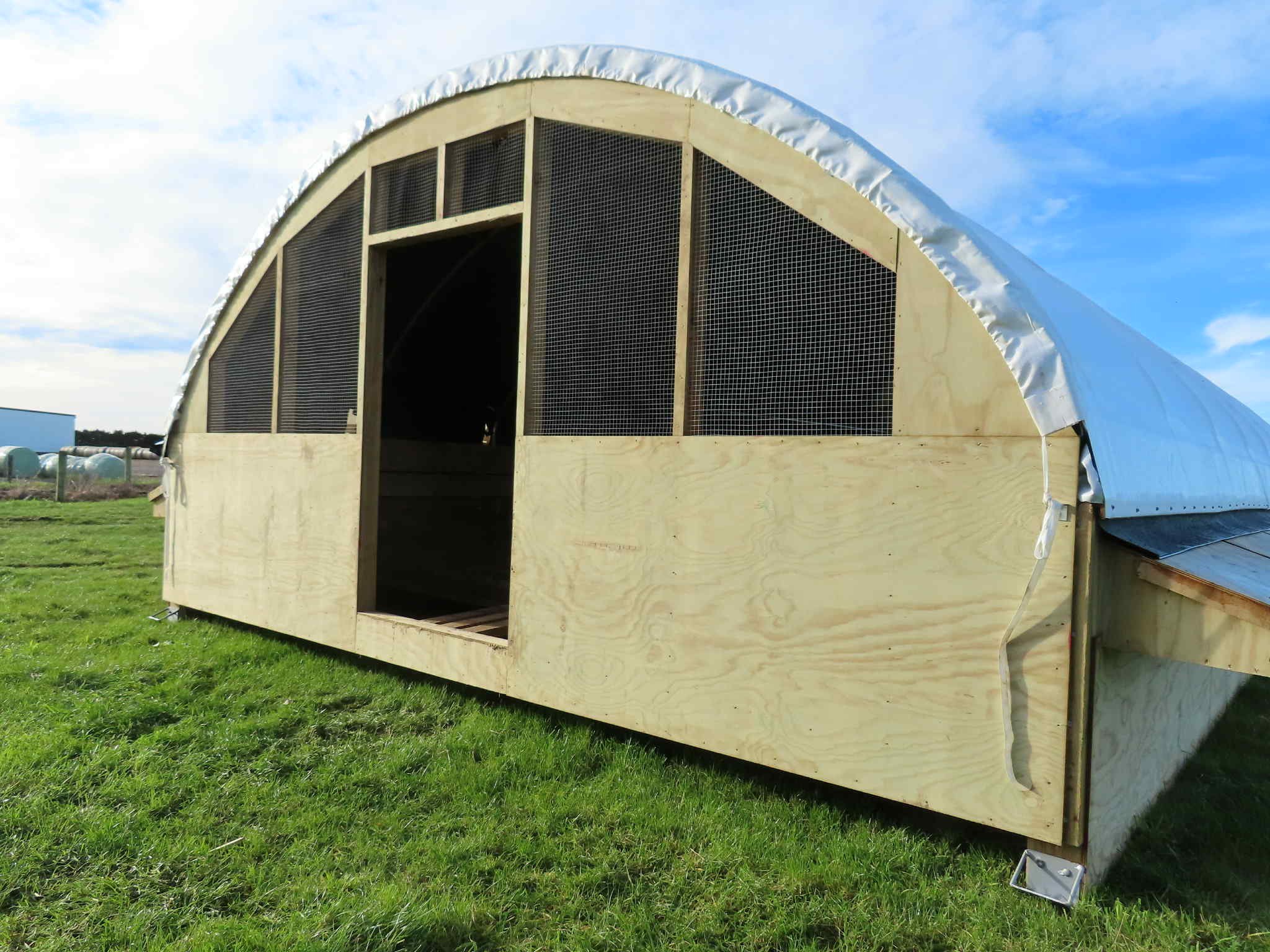 Unfinished wooden greenhouse with a curved metal roof on a grassy field, with a blue sky in the background.