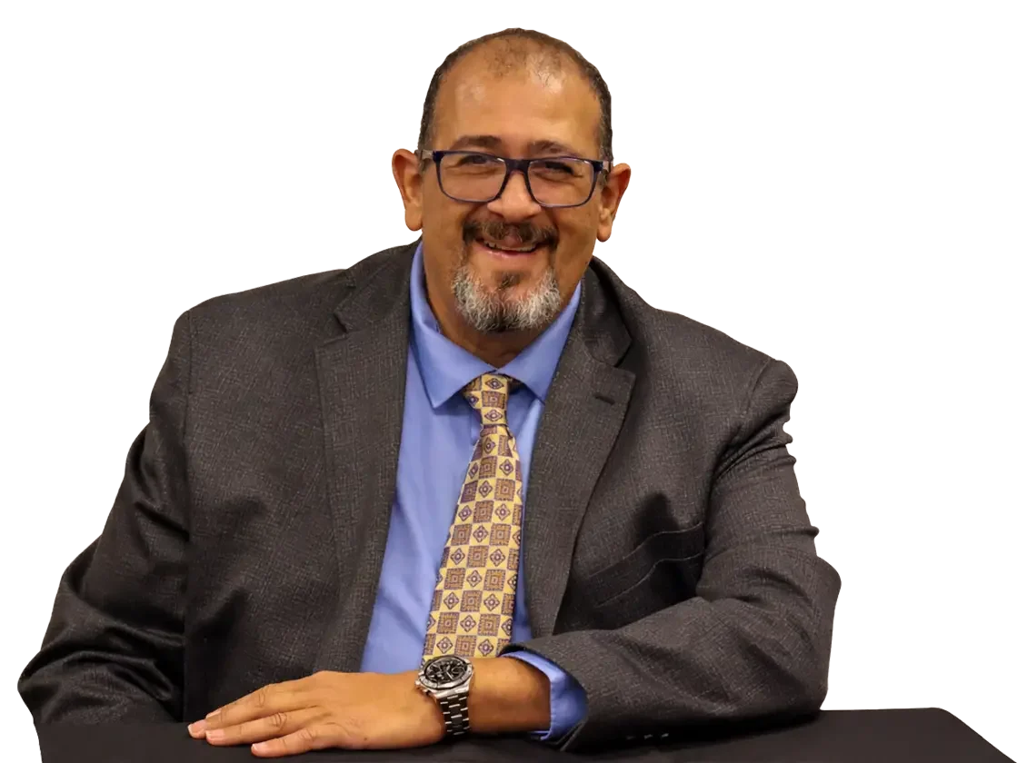 portrait of a middle-aged man wearing glasses, a dark gray suit, light blue shirt, patterned yellow tie, smiling, sitting at a table.