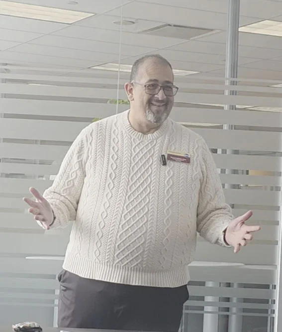 A man with glasses and a beard, wearing a cream cable-knit sweater, standing in an office with glass walls and a striped pattern, smiling with his hands raised.