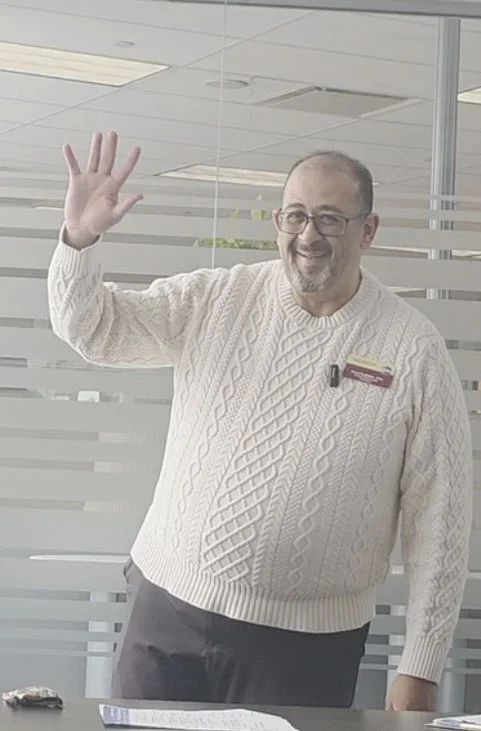 A man with glasses and a beard, wearing a cream cable-knit sweater, is standing and waving inside an office with glass walls and horizontal striped patterns.