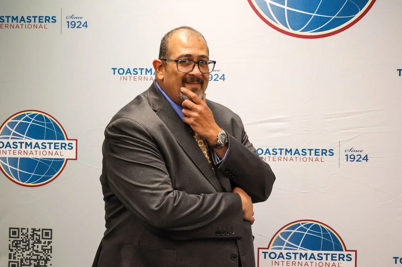 A man in a gray suit and glasses, with a watch, posing with his hand on his chin in front of a Toastmasters International banner.