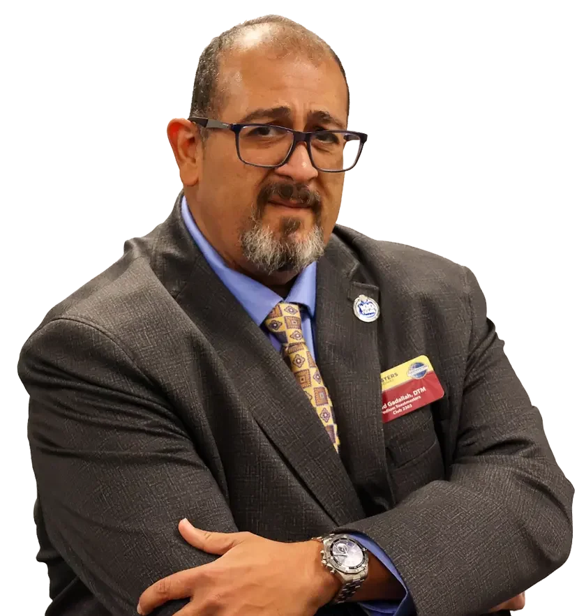 A man with glasses, facial hair, and a receding hairline in a suit and tie, crossing his arms, standing in front of a plain background.