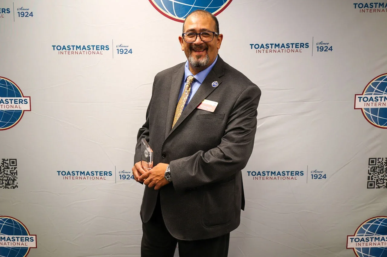 A man in a suit holding an award stands in front of a Toastmasters International backdrop, with logos and QR codes.