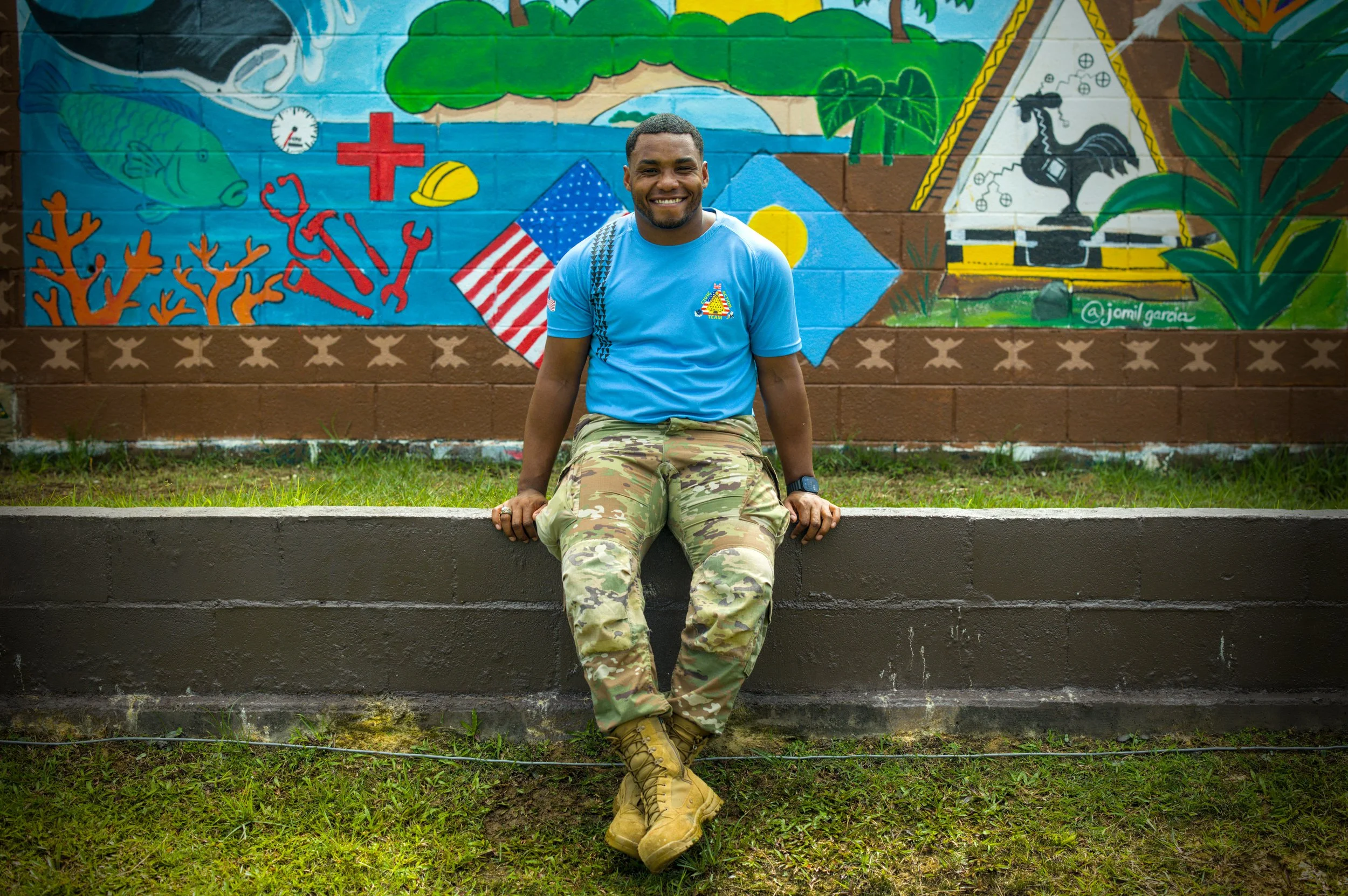 A young man with short blond hair smiling outdoors, wearing a dark green t-shirt with a colorful logo on the chest and a small flag on the sleeve.