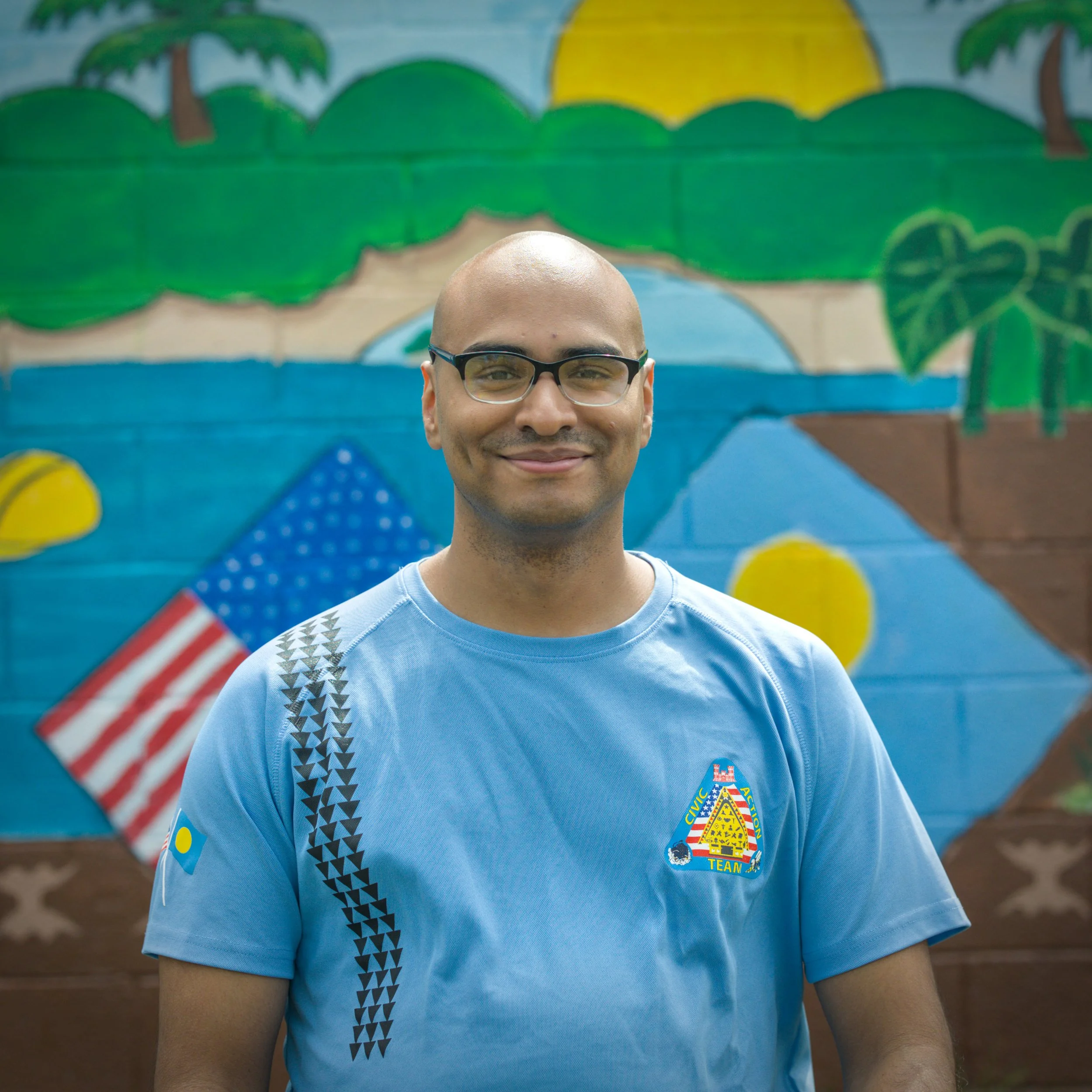 A smiling man in U.S. Army camouflage uniform standing against a beige textured wall.