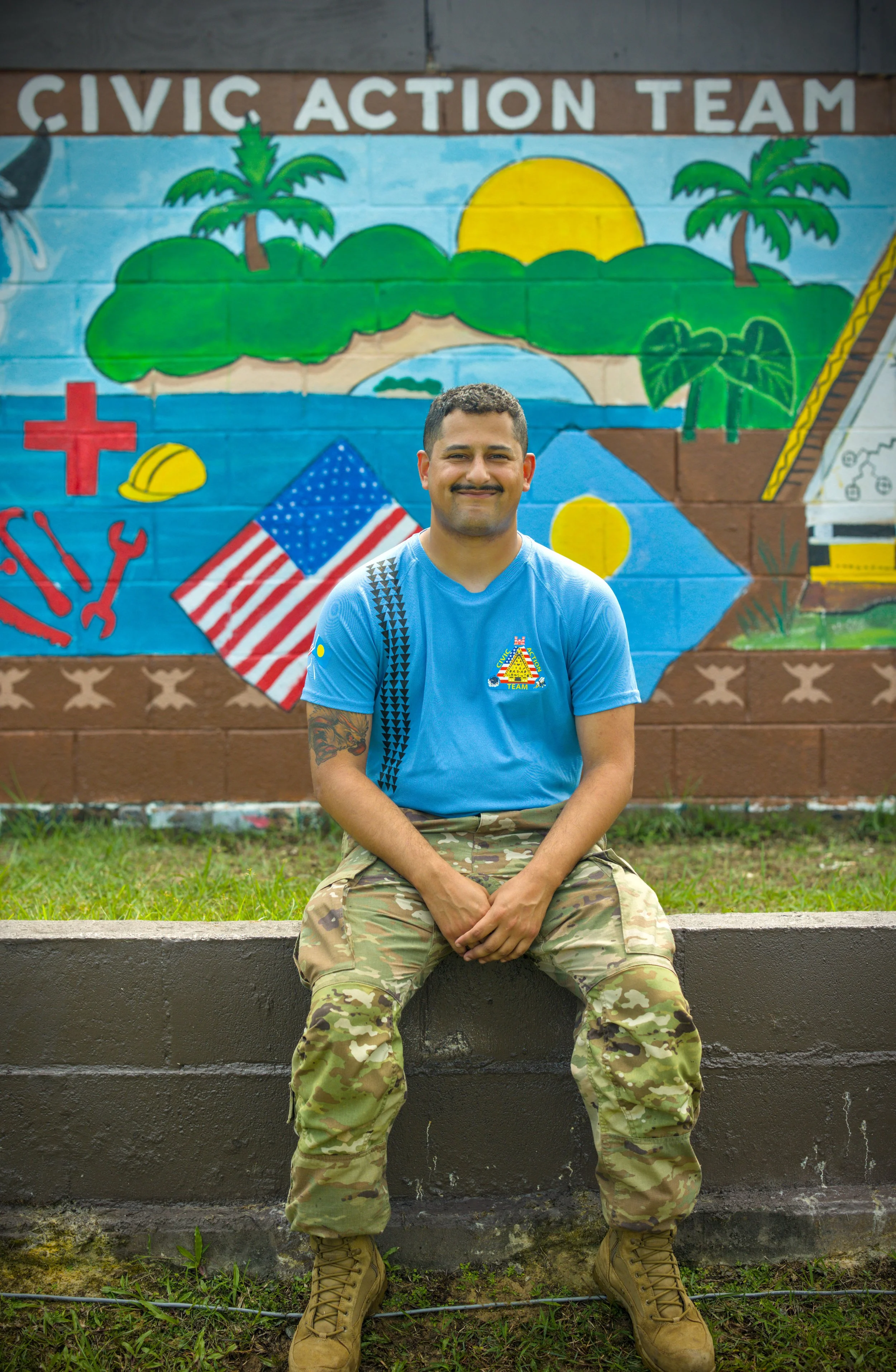 A man dressed in U.S. Army camouflage uniform standing against a beige wall, smiling at the camera.