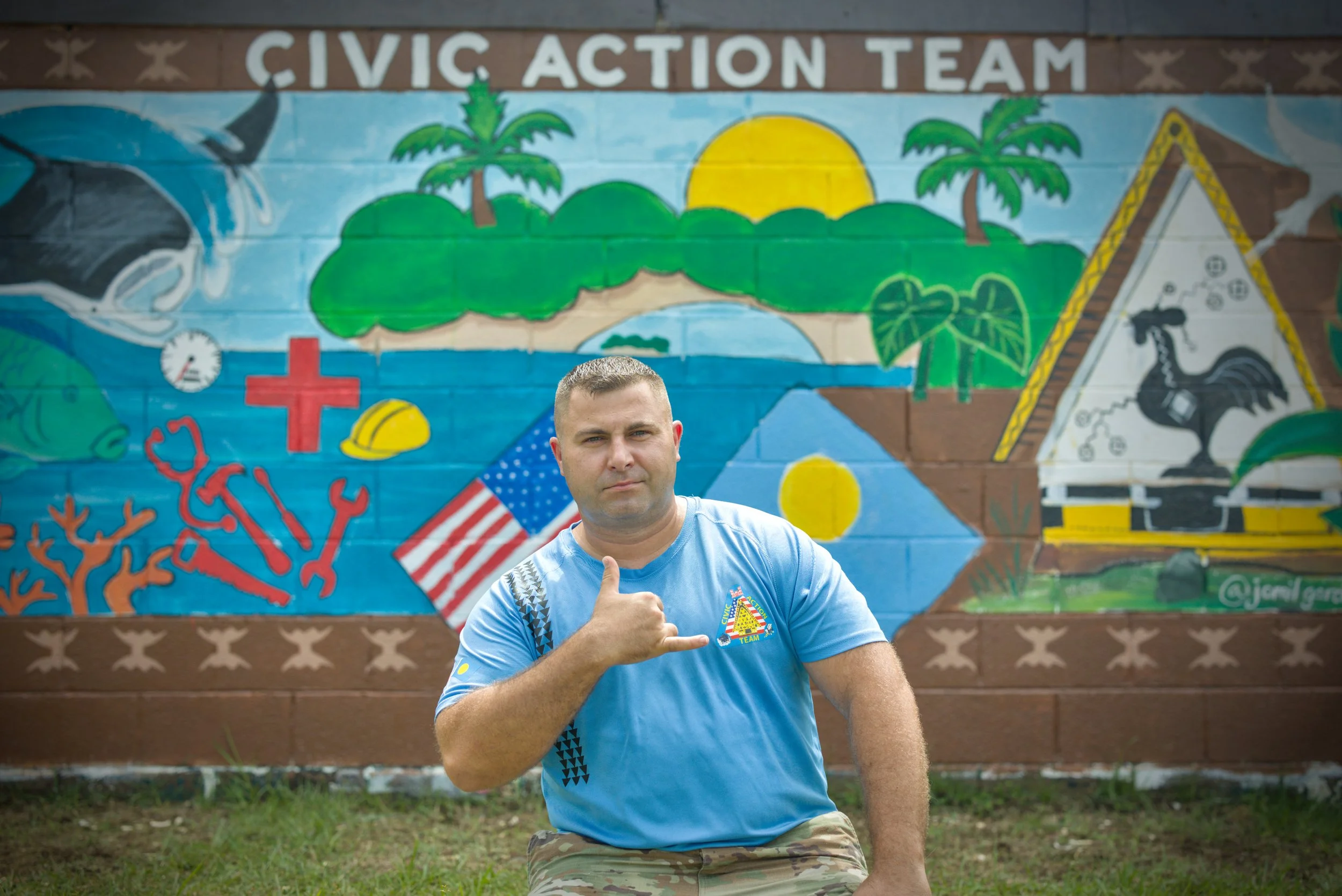 Portrait of a smiling young man in military camouflage uniform standing against a beige wall.