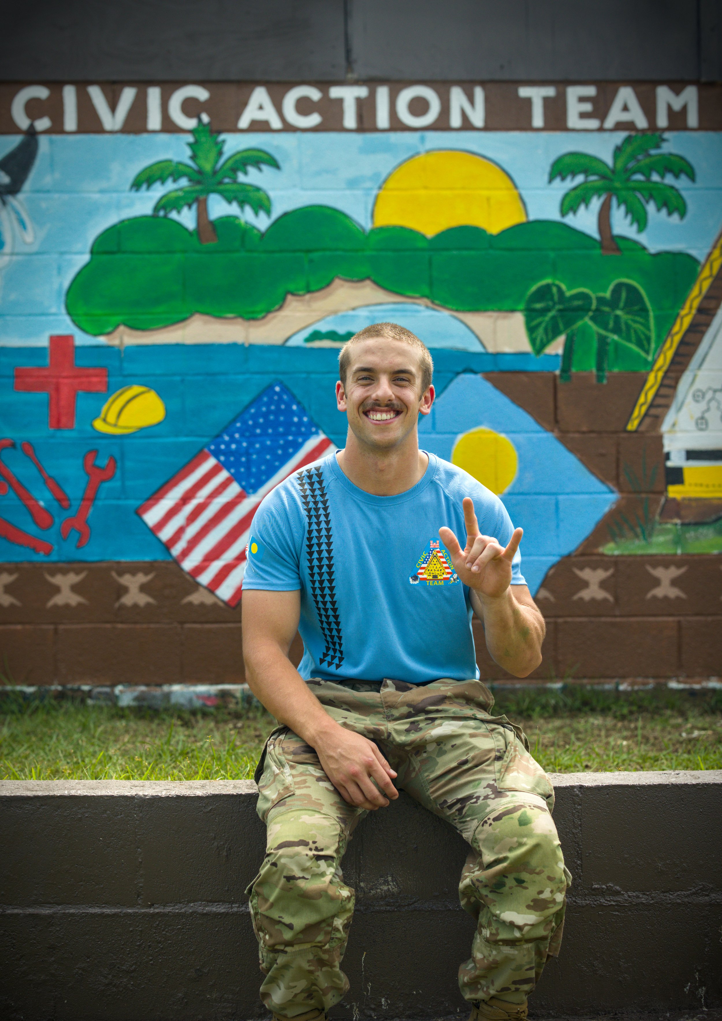 A young man in U.S. Army camouflage uniform standing against a beige wall.